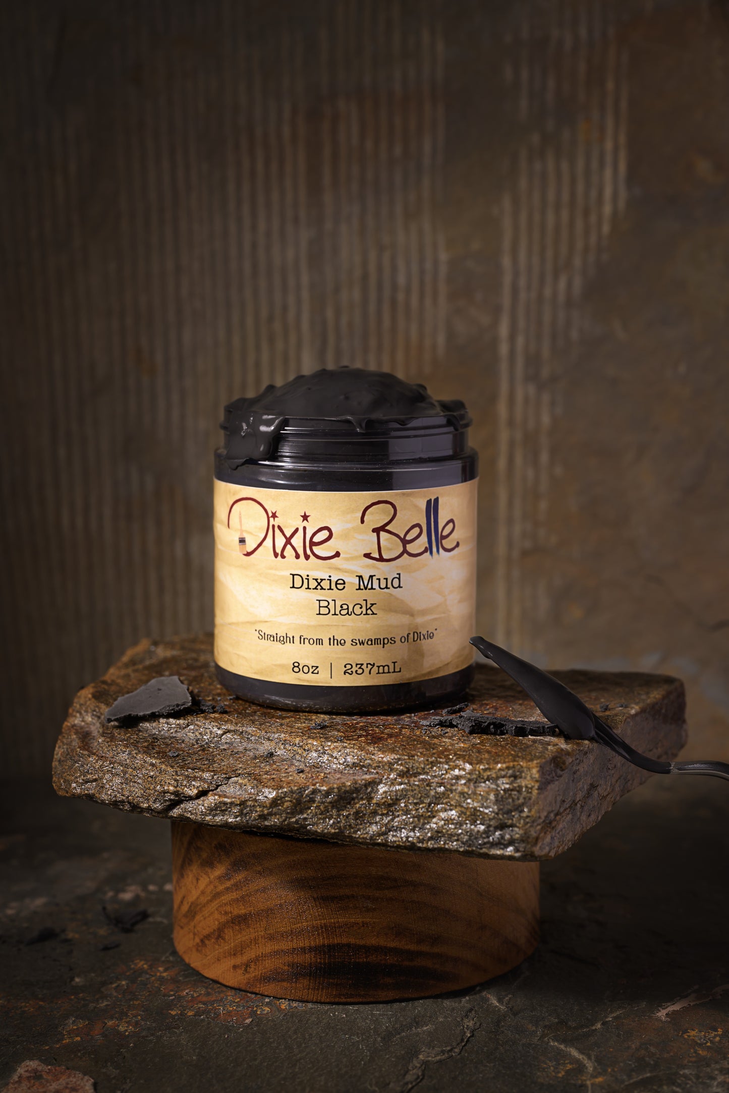 A jar of Dixie Belle Dixie Mud Black, 8 oz (237 mL), sits on a rough stone slab, which is supported by a round wooden base, against a rustic brown background.