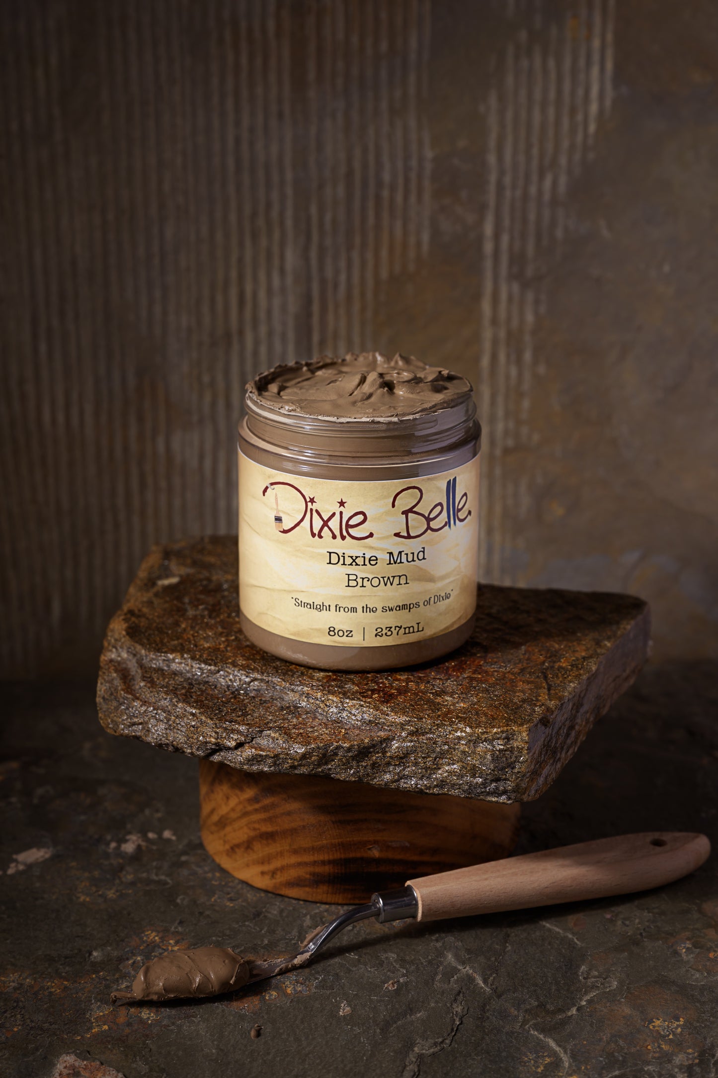 A jar of Dixie Belle Dixie Mud Brown sits on a stone slab and wooden base. The label reads "Straight from the swamps of Dixie" and indicates the container holds 8oz (237mL) of product.