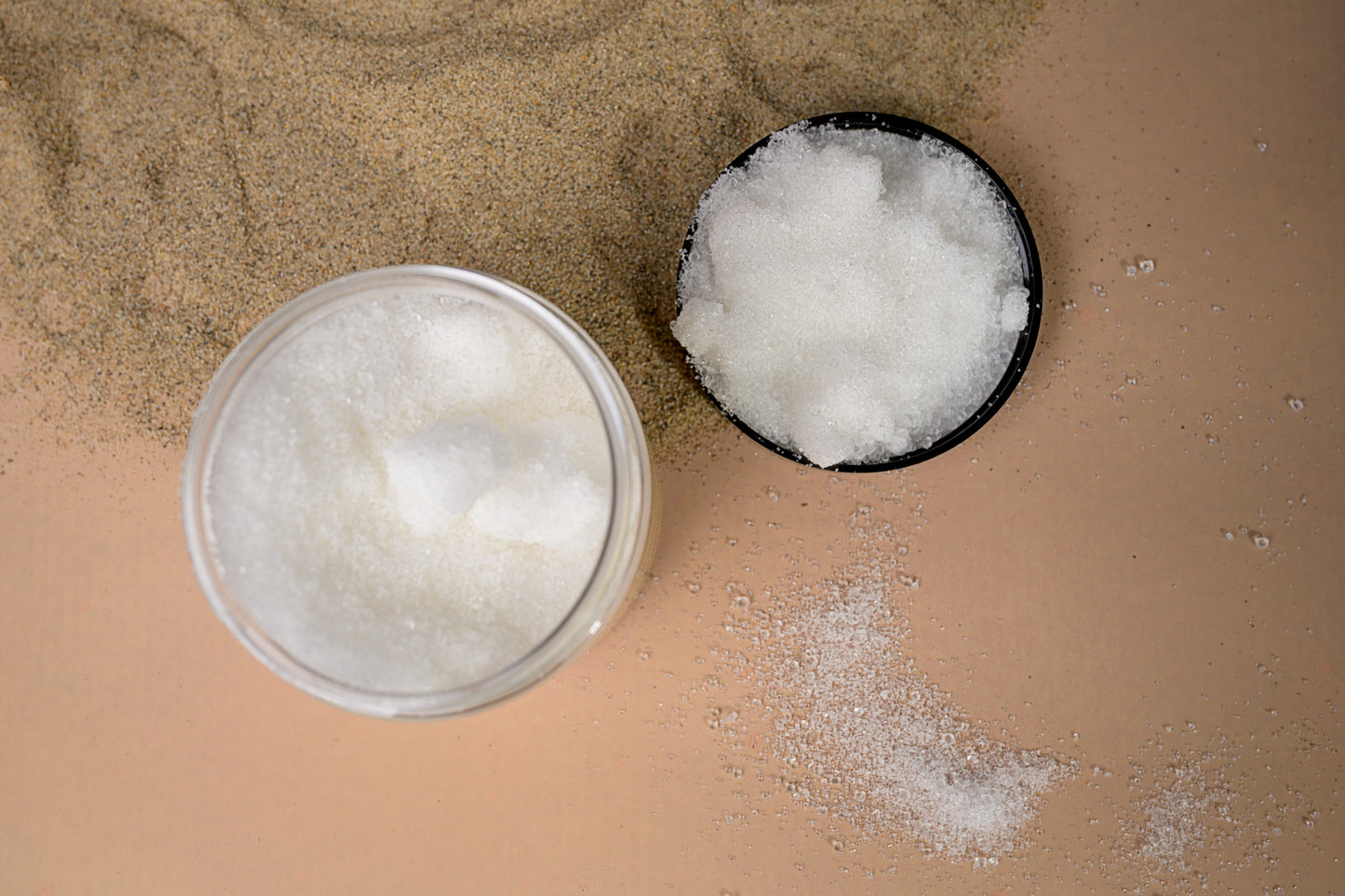 Top-down view of a glass jar and a black lid, both filled with whitish clear granules, placed on a beige surface sprinkled with sand and white granules.