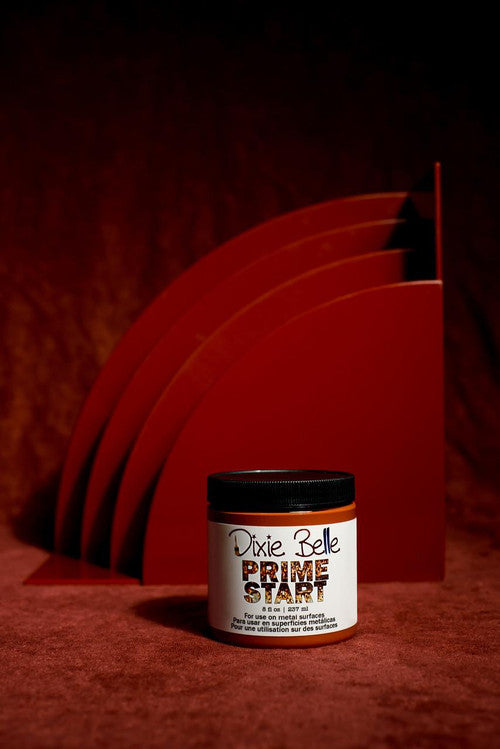 A jar of Dixie Belle Prime Start stands on a red surface with abstract, curved red panels in the background, all under dramatic lighting.
