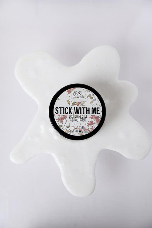 A container of “Stick With Me” Dixie Shine glue sits on a white surface, surrounded by a splatter-shaped spill of glue. The round label features decorative floral accents.
