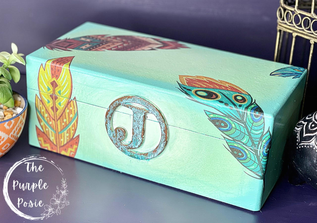 Belles and Whistles Feathers - Rice Decoupage Paper
Here is a a plain wooden box painted in a light green and has an iron, patinaed "J" with a metal circle around the "J" on the front of the box. The box can be opened like a jewelry box. It has some of the Belles and Whistles decoupage feathers used on the box. It is staged on a purple surface with the same purple behind the box. There is a yellow planter with a green plant and white pebbles in front of the left side of the box.