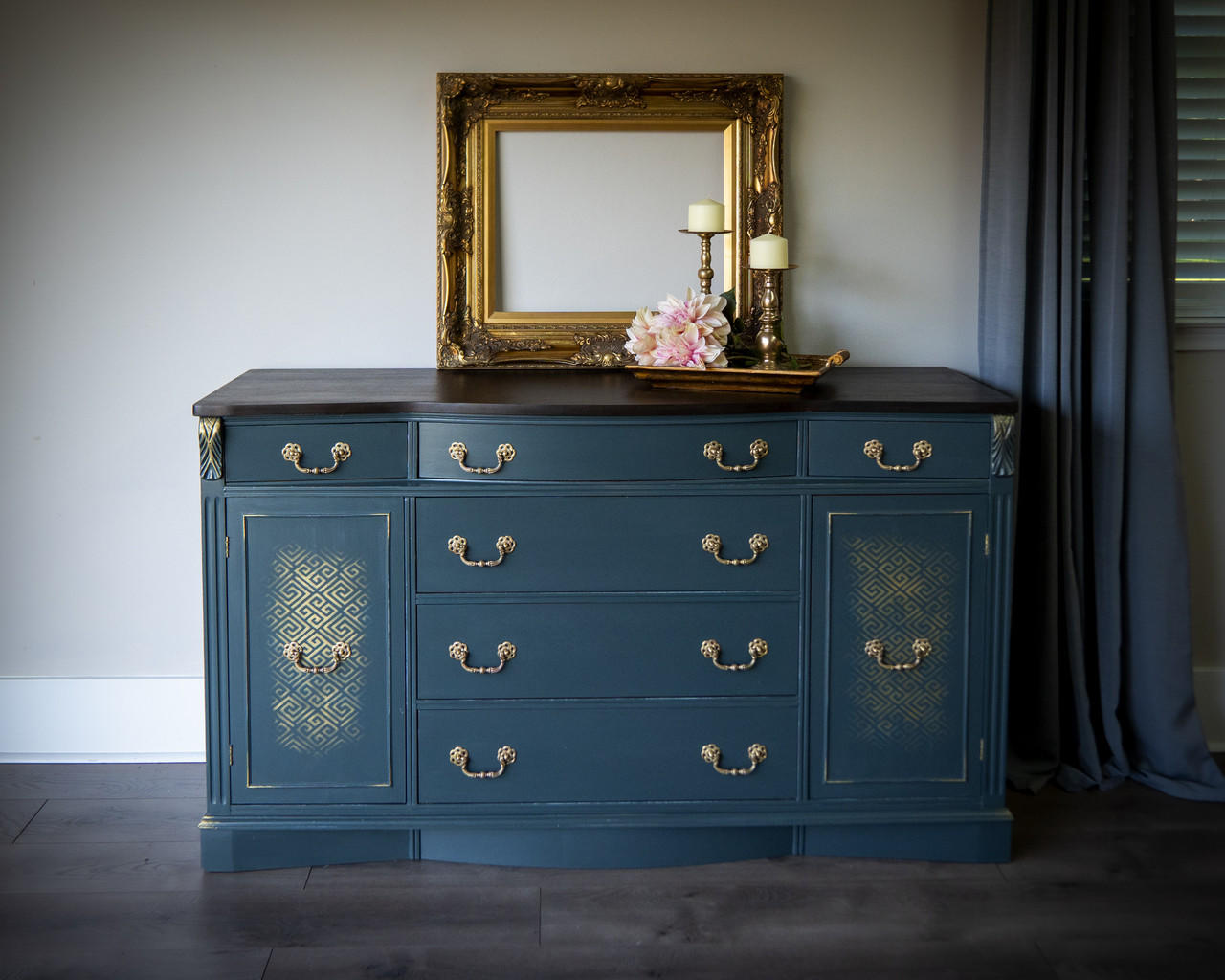 Belles and Whistles Greek Key - Stencil provides an extra decorative touch to any project! Dresser painted in a muted navy blue with gold hardwares and Greek Key - Stencil in gold faded a bit on the doors of the piece. Staged with a gold frame on the center of the dresser , with gold tray and two candle holder on top with some flowers.