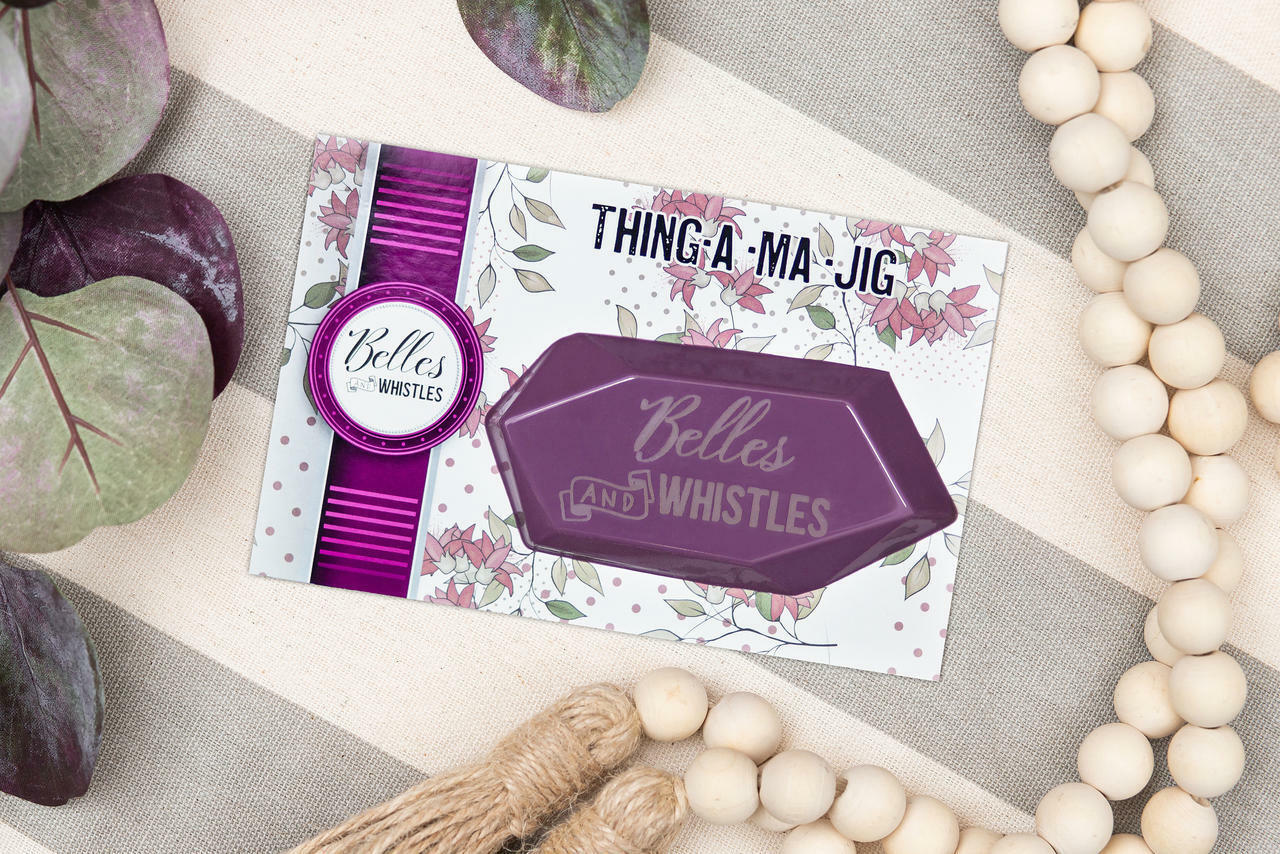 Belles and Whistles Thing-a-ma-jig - Thing-a-ma-jig in it's packaging sitting on a gray and ivory striped table with ivory beads and green & purple leaves around.