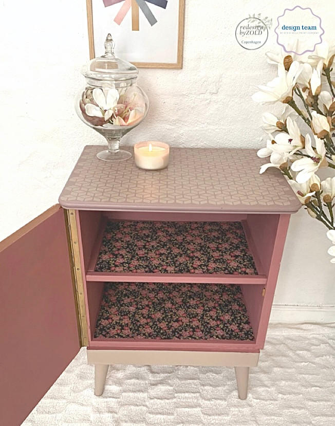 Belles and Whistles Trippy Blocks - Stencil. A pink chalk painted night stand with the front door open. The inside shelves are covered with pink and black floral paper. the top of the nightstand has the Trippy Blocks stencil applied in tan chalk paint. The base and legs of the table are painted with tan chalk paint. Staged on white carpet, in front of a white wall. White flowers are to the right of the night stand, and a glass vase holding flowers and a white candle sit on top of the night stand.