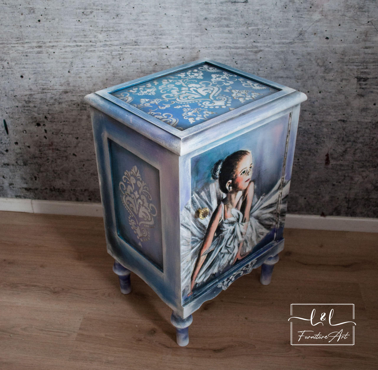Belles and Whistles Victorian Damask - Stencil provides an extra decorative touch to any project! A small blue and white chalk painted night stand with a picture of a young girl in a white dress applied as a decoupage to the front. The Victorian Damask stencil is applied to the top and side with white Chalk Paint. Staged on a brown wooden floor, in front of a gray wall with white wooden trim.