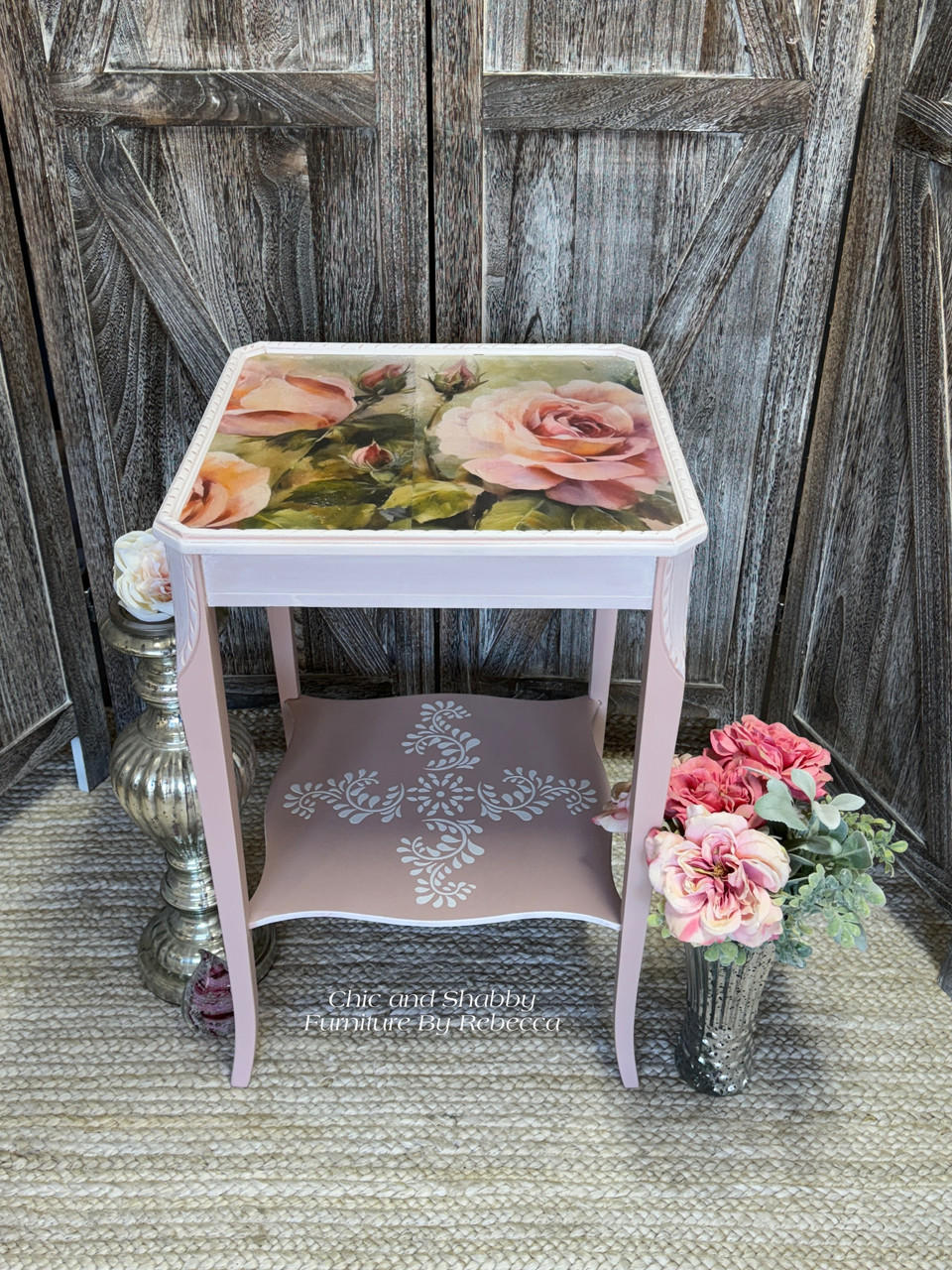 A vintage, shabby-chic style side table with floral motifs, painted in pastel pink and cream colors, elegantly standing against a painted rustic wooden backdrop with a bouquet of artificial flowers made using Blushing Roses A1 Rice Decoupage Paper.