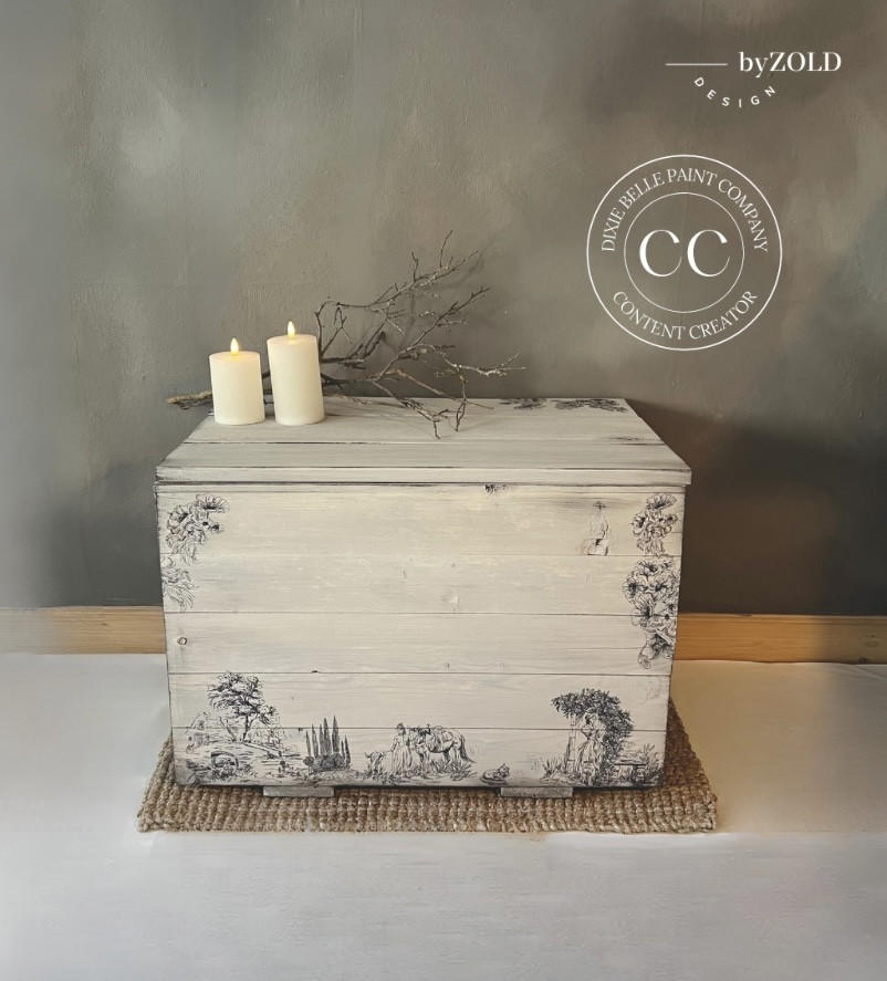 An off-white painted wooden chest with intricate black illustrations from the Cottage Toile Transfer, placed against a gray wall. The chest has two lit small white candles and a small branch on top. A jute mat lies underneath it. A logo for "Dixie Belle Paint Company Content Creator" is seen on the picture.