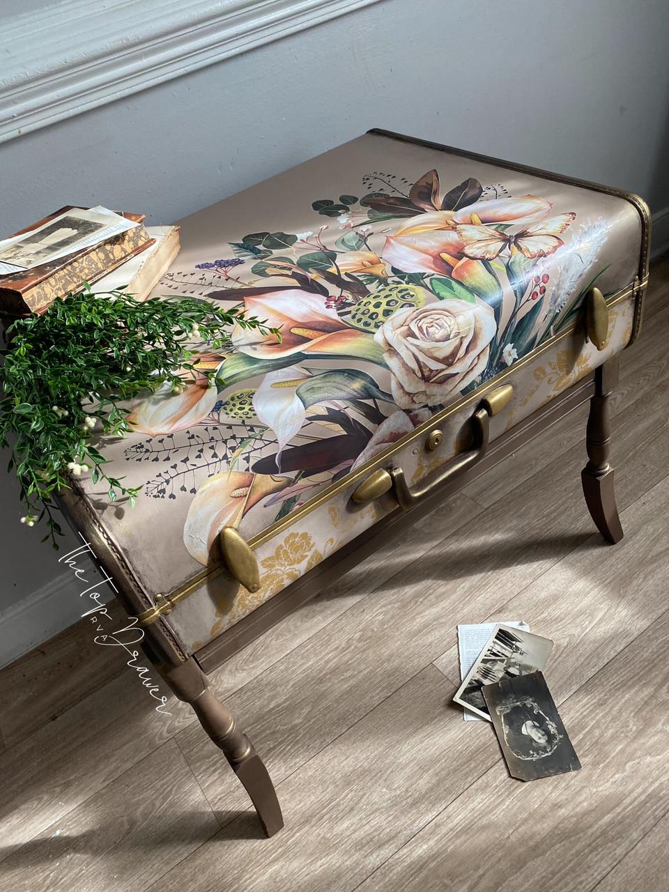 A vintage suitcase repurposed as a bench, adorned with floral artwork on top. A small plant and stacked books sit on the bench, with old photographs scattered on the wooden floor nearby.