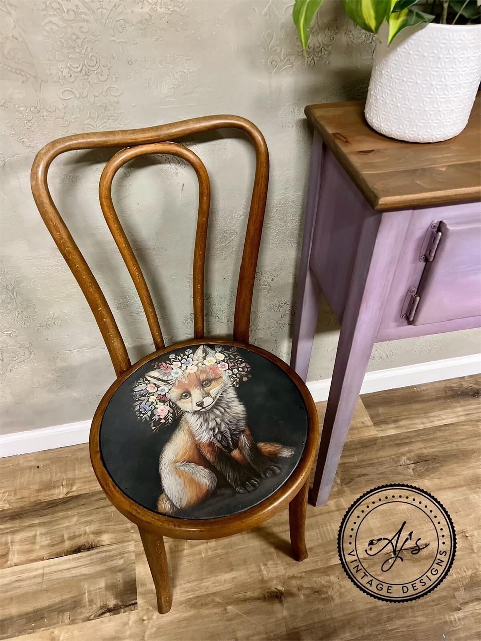 A wooden chair with a circular seat cushion featuring a painted fox wearing a floral crown. The chair is positioned on a wooden floor beside a small purple-painted table with a potted plant on top. The wall behind is textured. An "AJ's Vintage Designs" watermark logo is on the floor.