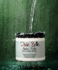 A jar of Dixie Belle Gator Hide is splashed with a stream of water against a green background, demonstrating its water-repellent properties. Water droplets are seen around and on the container.