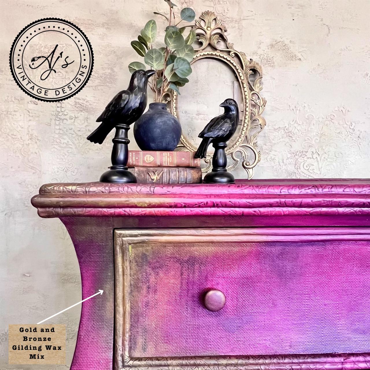 A vintage dresser with a gradient of purple and gold hues is adorned with two black bird figurines, a small vase with leaves, and stacked books. An ornate mirror is placed behind them. The image showcases the mix of Gold and Bronze Gilding wax.