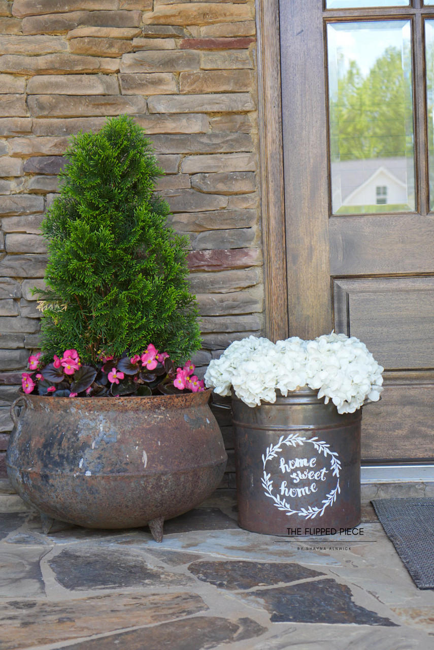 A charming entrance with a rustic metal pot and a decorative bucket, filled with pink and white flowers respectively, beside a topiary on a stone-clad porch. A reflection of a house is visible in the door’s window.