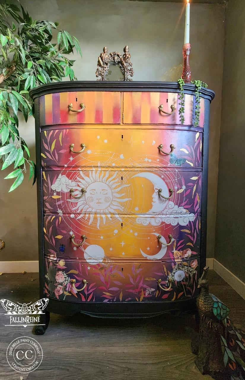 A colorful wooden dresser painted with a celestial theme, featuring a sun, moon, and stars. The dresser has ornate handles, sits in a room with leafy plants, and a single candle rests on top.
