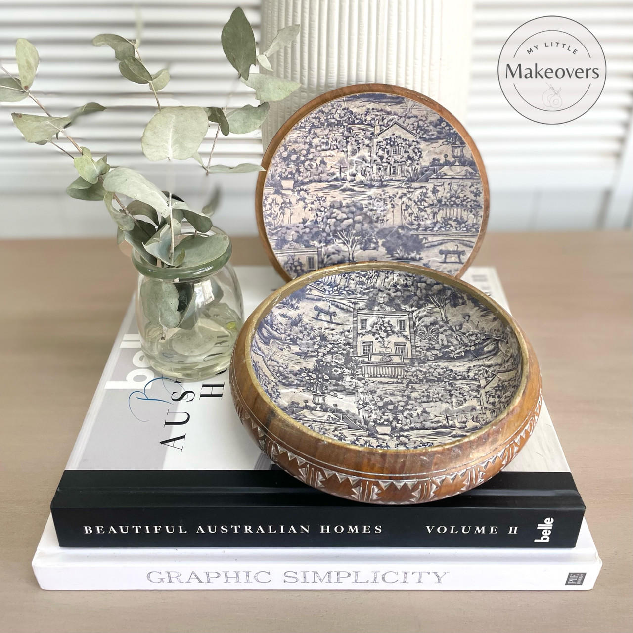 A decorative open box with intricate blue and white patterns is displayed on top of a stack of books. A small glass jar with green leaves is placed beside the box. The books are titled "Beautiful Australian Homes," and "Graphic Simplicity."