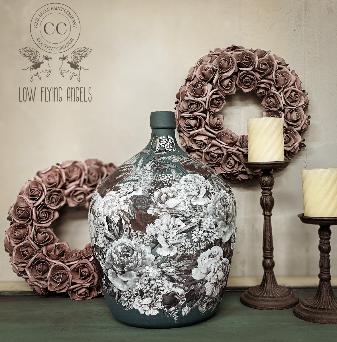 A large, intricately decorated ceramic floral vase is centered against a light wall. It is flanked by two metal candlesticks with candles and adorned with two wreaths made of dusty rose-colored flowers. The backdrop features a logo for "Low Flying Angels."