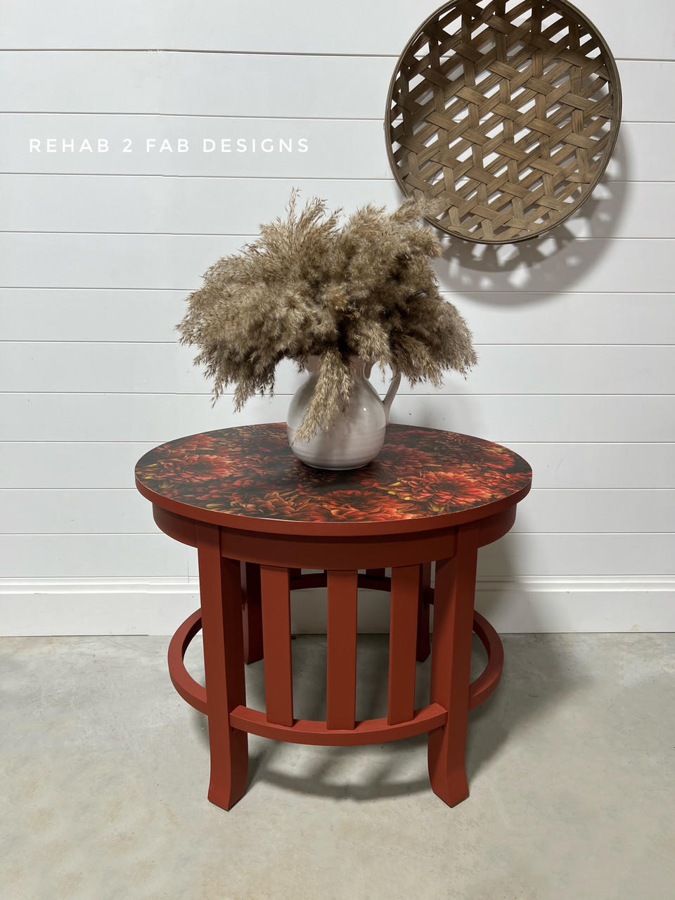 A round wooden end table with a red and black patterned top using the Moody Mums A1 Rice Decoupage Paper. Adorned with a white vase filled with fluffy pampas grass, set against a white shiplap wall with a decorative round wooden tray hanging.