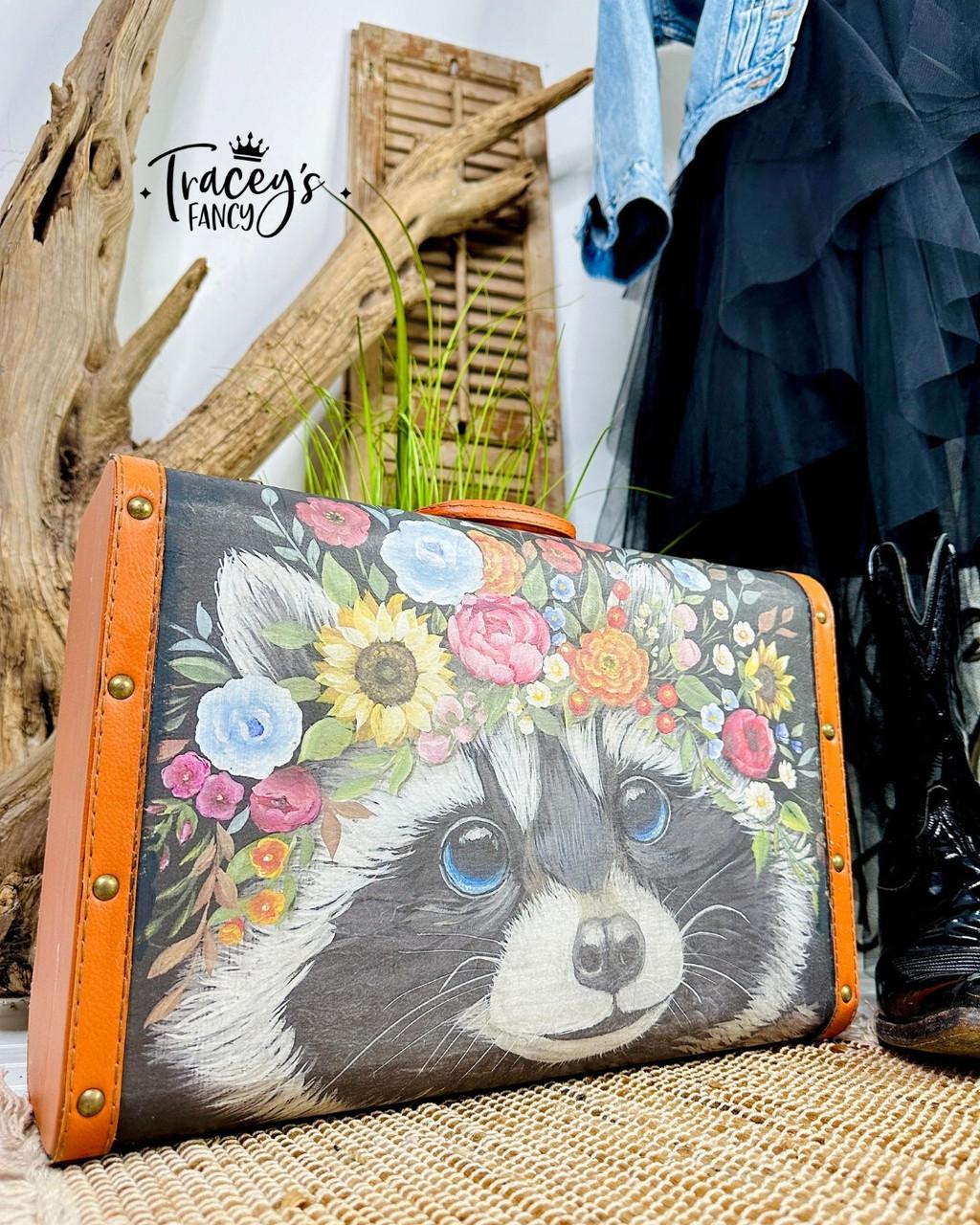A vintage suitcase with an orange border features a painted raccoon wearing a colorful flower crown. The suitcase is positioned on a woven surface with a rustic wood backdrop, green plants, cowboy boots, and a denim jacket draped nearby. The watermark logo "Tracey's Fancy" is visible in the background.