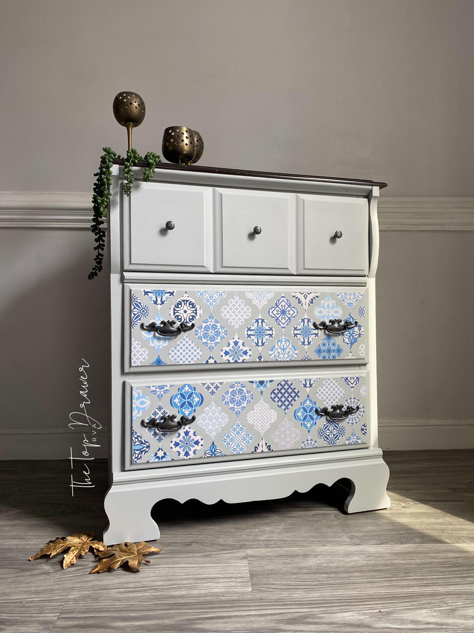 A gray 3 drawer dresser. Sapphire Sonata Transfer is seen applied to the bottom two drawers, the bottom two drawers have long intricate hardware, the top drawer has knobs hardware. Staged with bronze cups with a vine on top of the piece and large gold leaves on the floor.