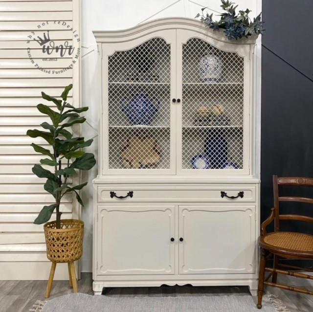 Endless Shore Silk All-In-One paint is perfect for painting kitchen cabinets, bathroom cabinets and painting furniture. White painted armoire with black handles. Styled next to a woven planter containing faux greenery. Wooden chair placed on the right of the armoire.