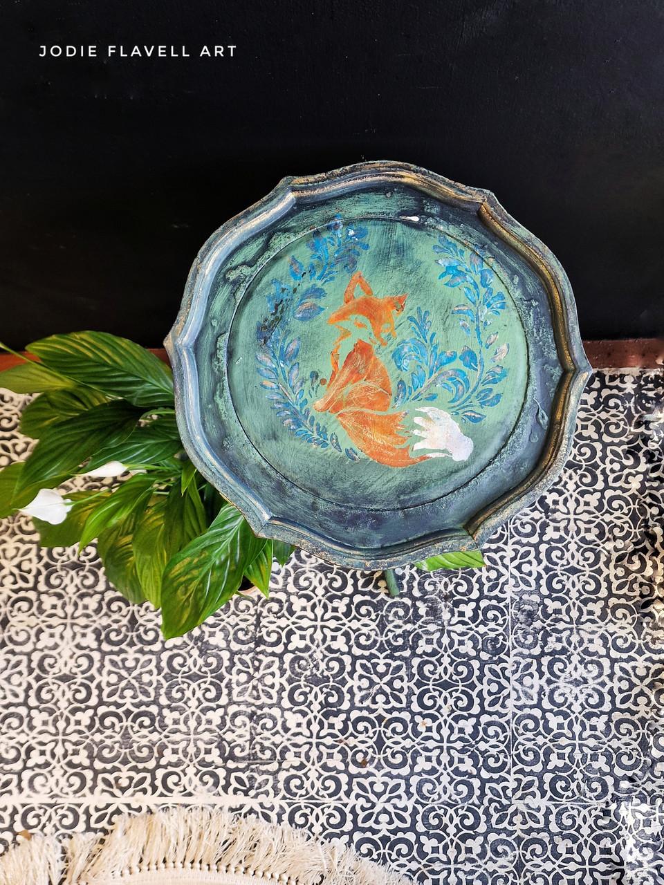 A round, intricately designed wooden tray features a painted image of a fox surrounded by blue floral patterns. The tray sits on a tiled surface with a plant nearby. Text in the top left corner reads "Jodie Flavell Art.