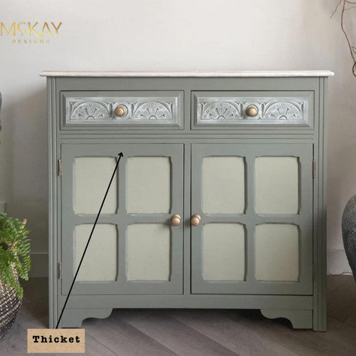 A light green cabinet with intricate patterns on the top drawers and gold knobs. "Thicket" is labeled with an arrow pointing to a section. The cabinet stands on a wooden floor with a potted plant nearby.
