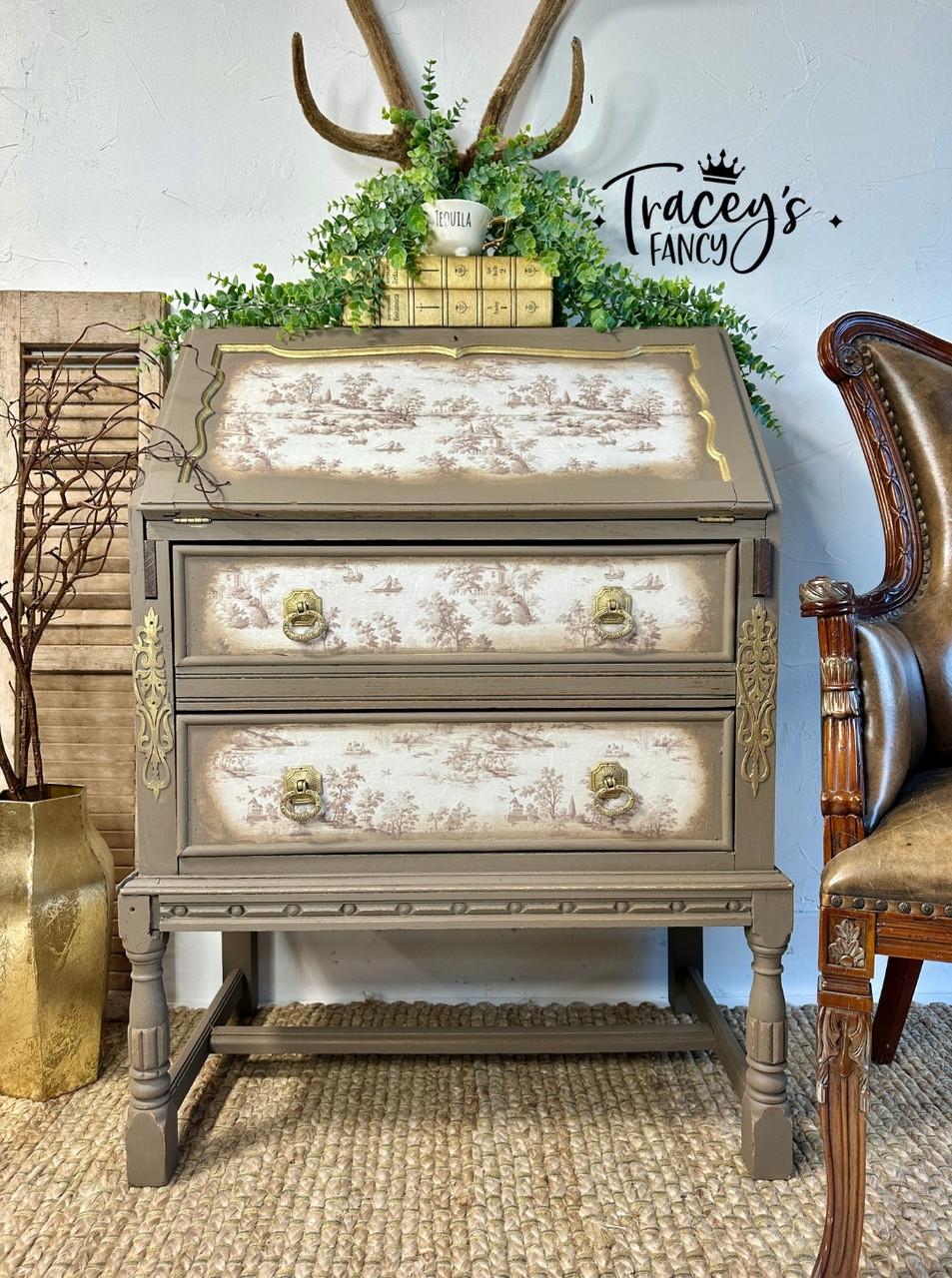 A vintage-style wooden chest with ornate gold details, floral-patterned drawers, greenery, and antlers on top sits against a white wall with "Tracey's Fancy" written on it. A brown chair is partially visible to the right.