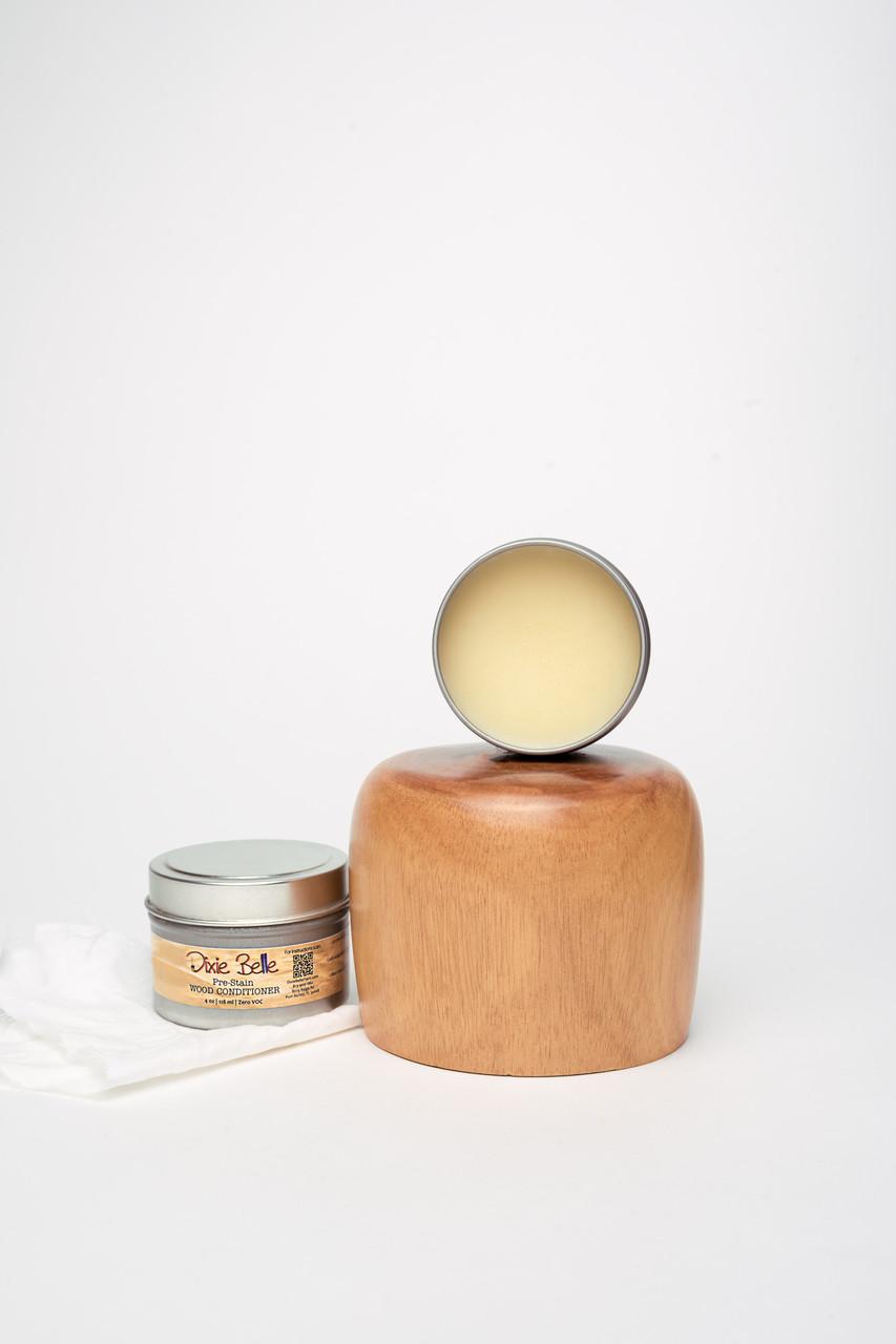 An open jar of Dixie Belle wood conditioner is placed in front of a wooden block and a polished wooden object. A white cloth rests beside the jar, all set against a plain white background.