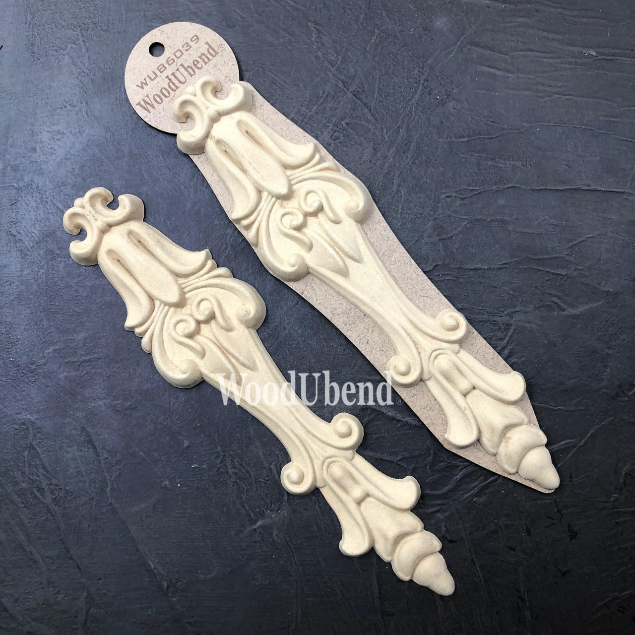 WoodUBend Applique #6039 (Set of 2) is the perfect mouldable embellishment for any project! This image shows Applique #6039 (Set of 2) with its packaging against a black background.