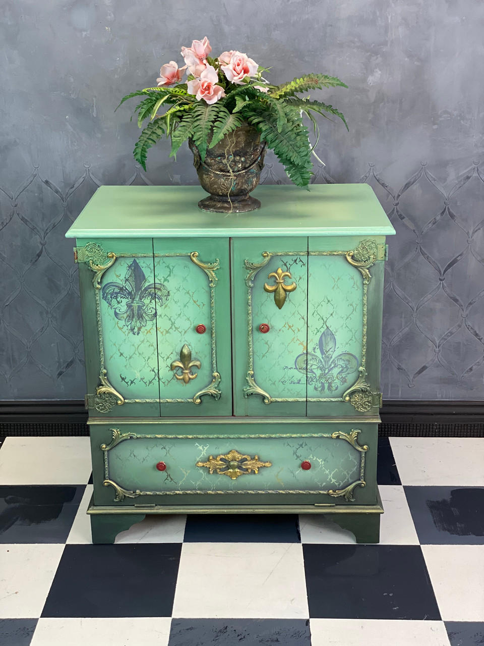 WoodUBend Fleur-de-lis Applique #X1011 (Set of 2) is the perfect mouldable embellishment for any project!
A cabinet table with Fleur-de-lis Applique #X1011 (Set of 2) on the cabinet doors opposite from each other. Staged with a rusted metal can with greenery and flowers inside on top of the table.