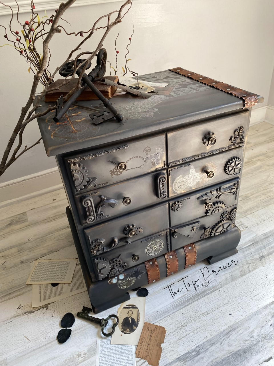 WoodUBend Trim #TR37 is the perfect mouldable embellishment for any project! A brown chalk painted dresser with brown metal strips, brown gears and WoodUBend attached to the front. Staged with large black keys and branches.