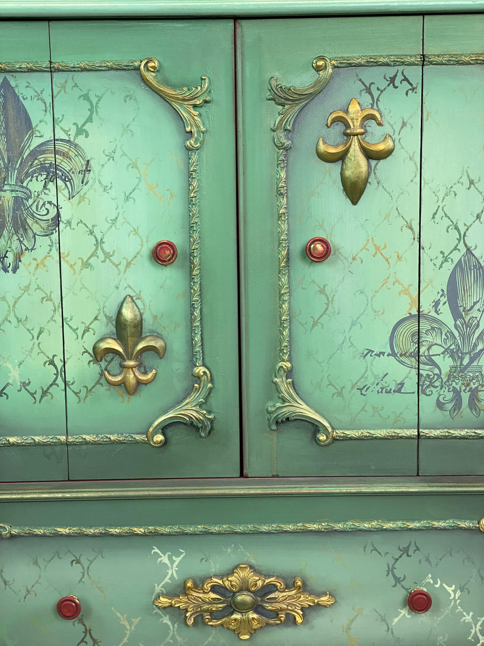 WoodUBend Trim #TR50a is the perfect mouldable embellishment for any project! A section of a green chalk painted cabinet that has several different WoodUBend moulds applied to it and accented in gold gilding wax. A Fleur-de-lis rub on transfer is applied to the doors, and a stencil pattern is chalk painted to the front of the piece in multiple chalk painted colors. The knobs are red.