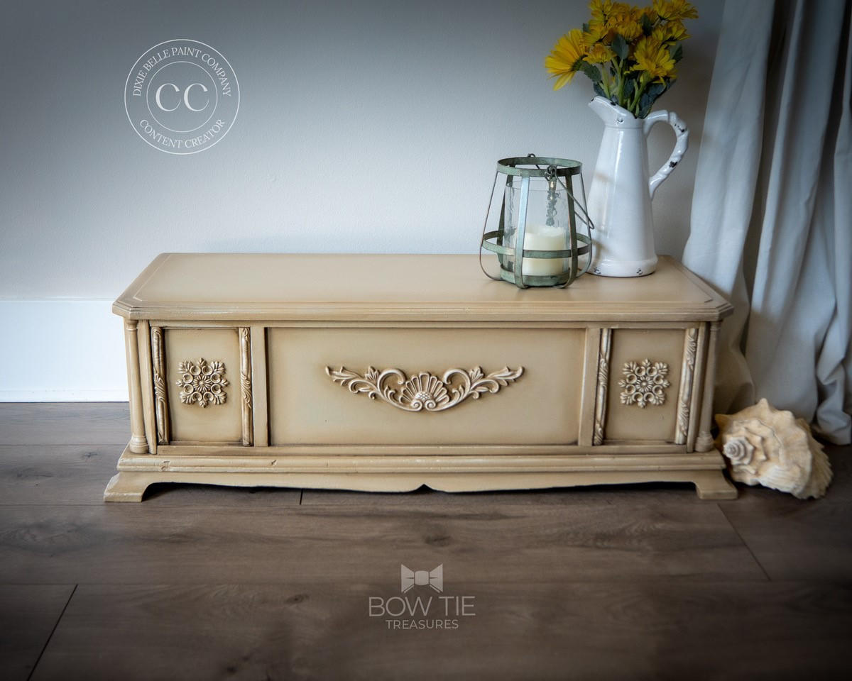 WoodUBend Trim #TR705 (Set of 2). A tan coffee table with a large WoodUBend scroll in the center front and a snowflake style WoodUBend piece on both end doors. WoodUBend strips line each of the doors. Staged on a wooden floor in front of a white wall with a white curtain to the right, a conch shell on the floor to the right. Sitting on the table is a white pitcher with yellow flowers and a glass candle holder.