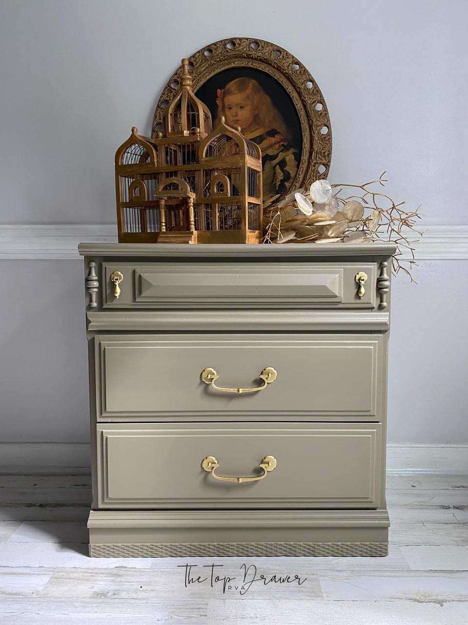 WoodUBend Trim #TR716 (Set of 2) is the perfect mouldable embellishment for any project! A brown chalk painted night stand with gold handles. WoodUbend is applied to the bottom front of the night stand. The night stand is staged on a whitewashed wood floor, in front of a gray wall with white wood trim. A brown wooden bird nest, brown branches and an oval picture of a young girl sit on top of the night stand.