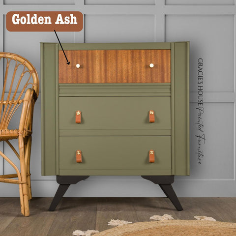 A small wooden cabinet with a modern design features three drawers. The top drawer is highlighted as “Golden Ash” wood. It stands on short black legs. A wicker chair is partially visible to the left. The wall paneling is light grey.
