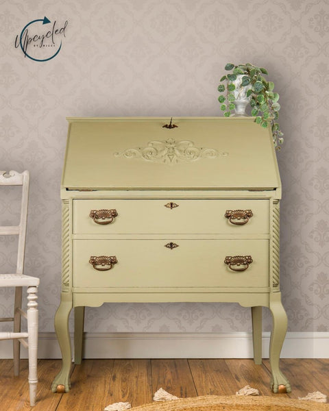 A vintage, pale green secretary desk with ornate details and brass handles stands against a patterned wallpaper. A potted trailing plant sits on top, and a white wooden chair is partially visible on the left.
