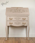 A vintage-style beige writing desk with ornate floral detailing, gold handles, curved legs, and a "Upcycled by Nicci" watermark on the wall behind it. The desk is placed on a wooden floor against a light wall.