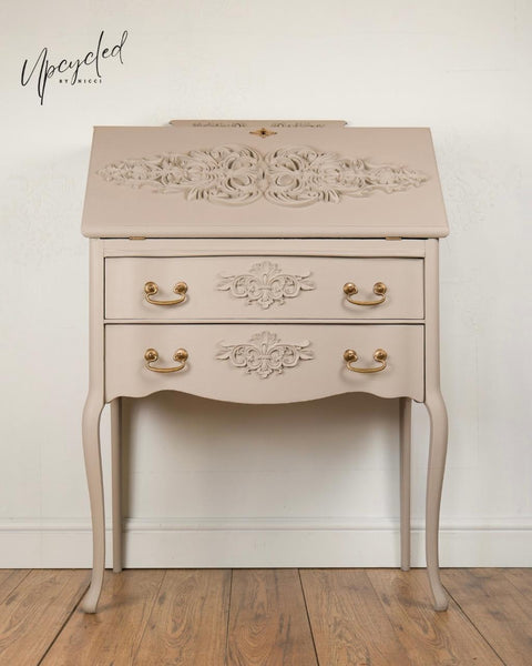 A vintage-style beige writing desk with ornate floral detailing, gold handles, curved legs, and a "Upcycled by Nicci" watermark on the wall behind it. The desk is placed on a wooden floor against a light wall.