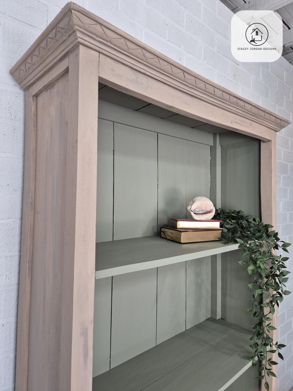 A wooden bookshelf with light brown trim and light green shelves holds a few books, a decorative shell, and a trailing green plant. The bookshelf stands against a white brick wall.