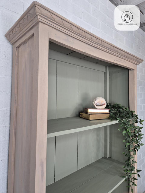 A wooden bookshelf with light brown trim and light green shelves holds a few books, a decorative shell, and a trailing green plant. The bookshelf stands against a white brick wall.