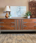 A mid-century modern wooden credenza with six drawers and vertical handles stands on a wood floor. It is topped with a lamp, potted plants, and decor, with a painting and ornate screen in the background. Watermark reads "Retro Revival, ABQ."