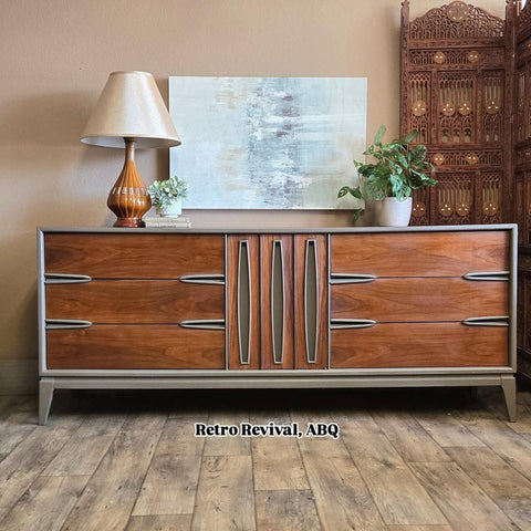 A mid-century modern wooden credenza with six drawers and vertical handles stands on a wood floor. It is topped with a lamp, potted plants, and decor, with a painting and ornate screen in the background. Watermark reads "Retro Revival, ABQ."