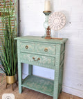 A small, decorative wooden table painted in light green with white floral patterns. It has two drawers with brass handles, a shelf, and a candle holder with a carved circular ornament on top. A tall green plant is beside it against a white brick wall.