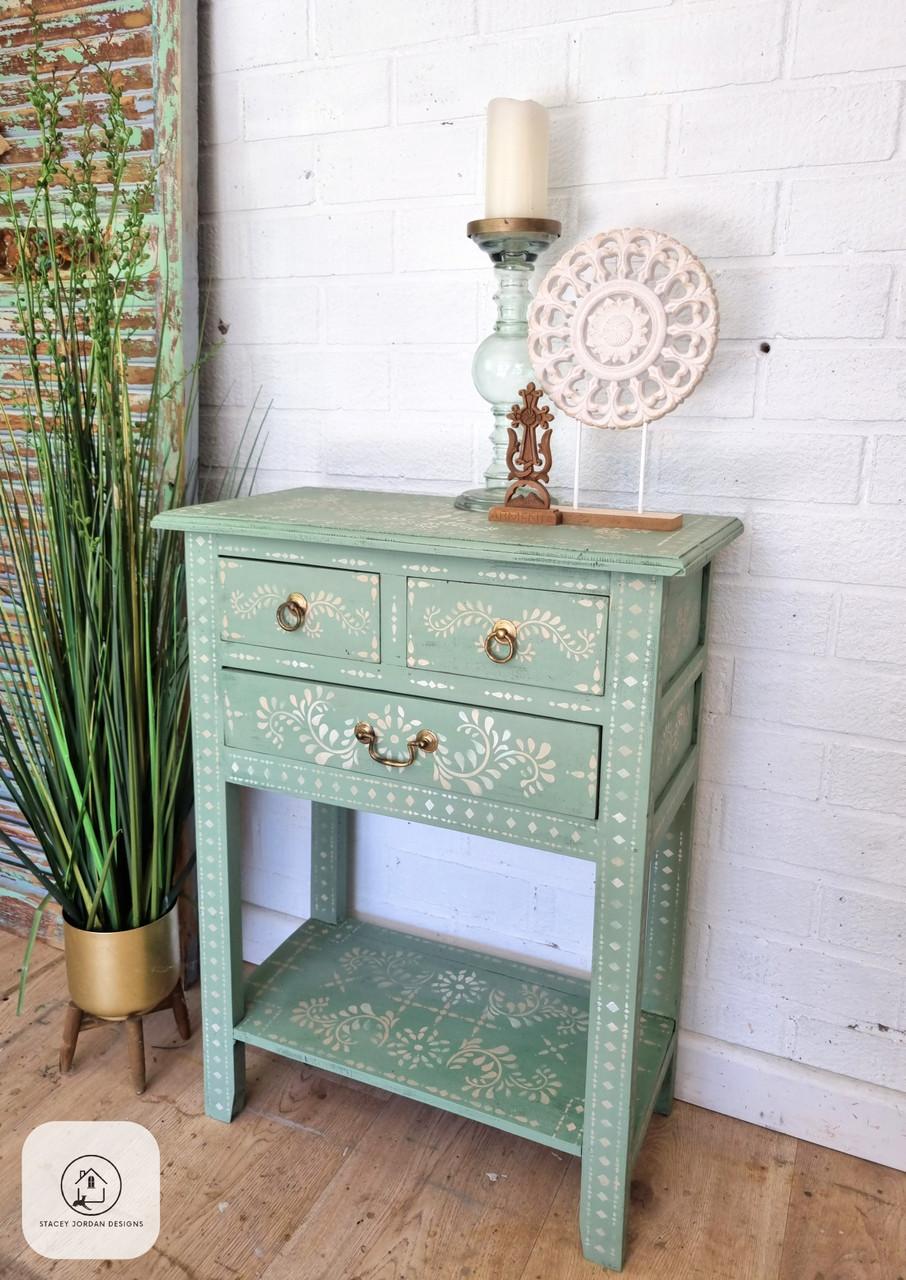 A small, decorative wooden table painted in light green with white floral patterns. It has two drawers with brass handles, a shelf, and a candle holder with a carved circular ornament on top. A tall green plant is beside it against a white brick wall.