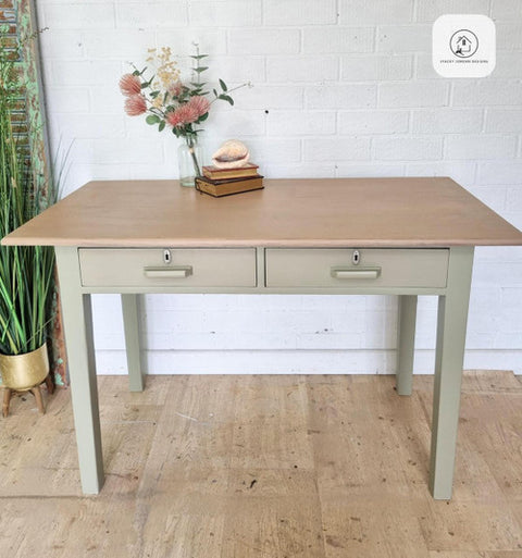 A wooden desk with two drawers stands on a light wood floor. On the desk are a vase with flowers, a stack of books, and a decorative shell. A tall plant is beside the desk against a white brick wall.