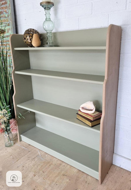 A light green wooden bookshelf with four shelves, holding a glass lamp, decorative items, and a few stacked books. The bookshelf stands on a wooden floor against a white brick wall, with flowers arranged nearby.