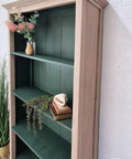 A wooden bookcase with green shelves holds a vase with flowers, a stack of books, a small decorative shell, and greenery, standing against a white brick wall.