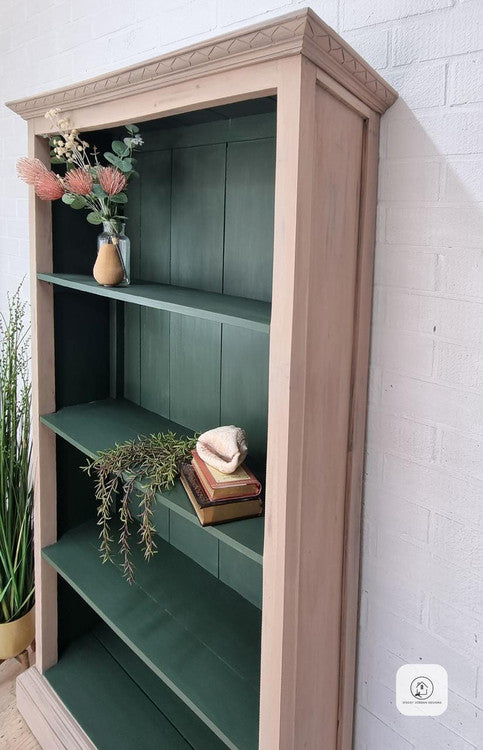 A wooden bookcase with green shelves holds a vase with flowers, a stack of books, a small decorative shell, and greenery, standing against a white brick wall.