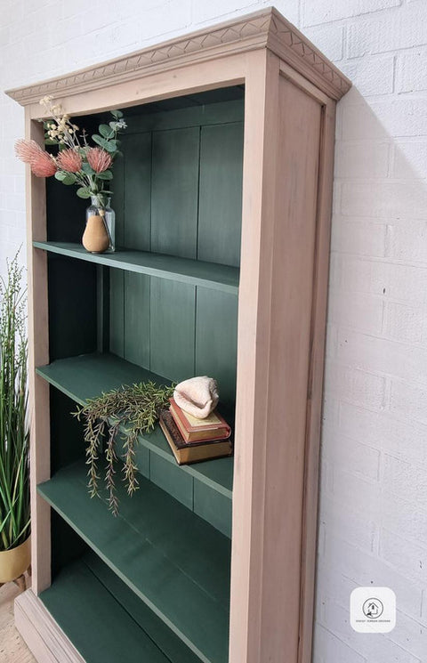 A wooden bookcase with green shelves holds a vase with flowers, a stack of books, a small decorative shell, and greenery, standing against a white brick wall.