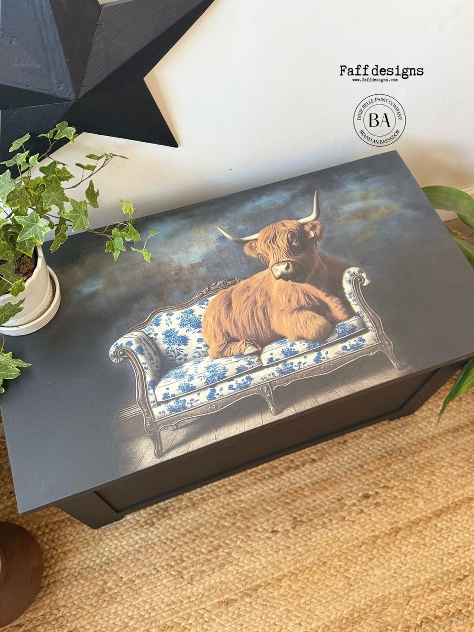 A painted chest features an image of a highland cow lounging on a blue and white floral sofa. A potted plant sits nearby on a woven rug, and a black star wall decoration is visible above.
